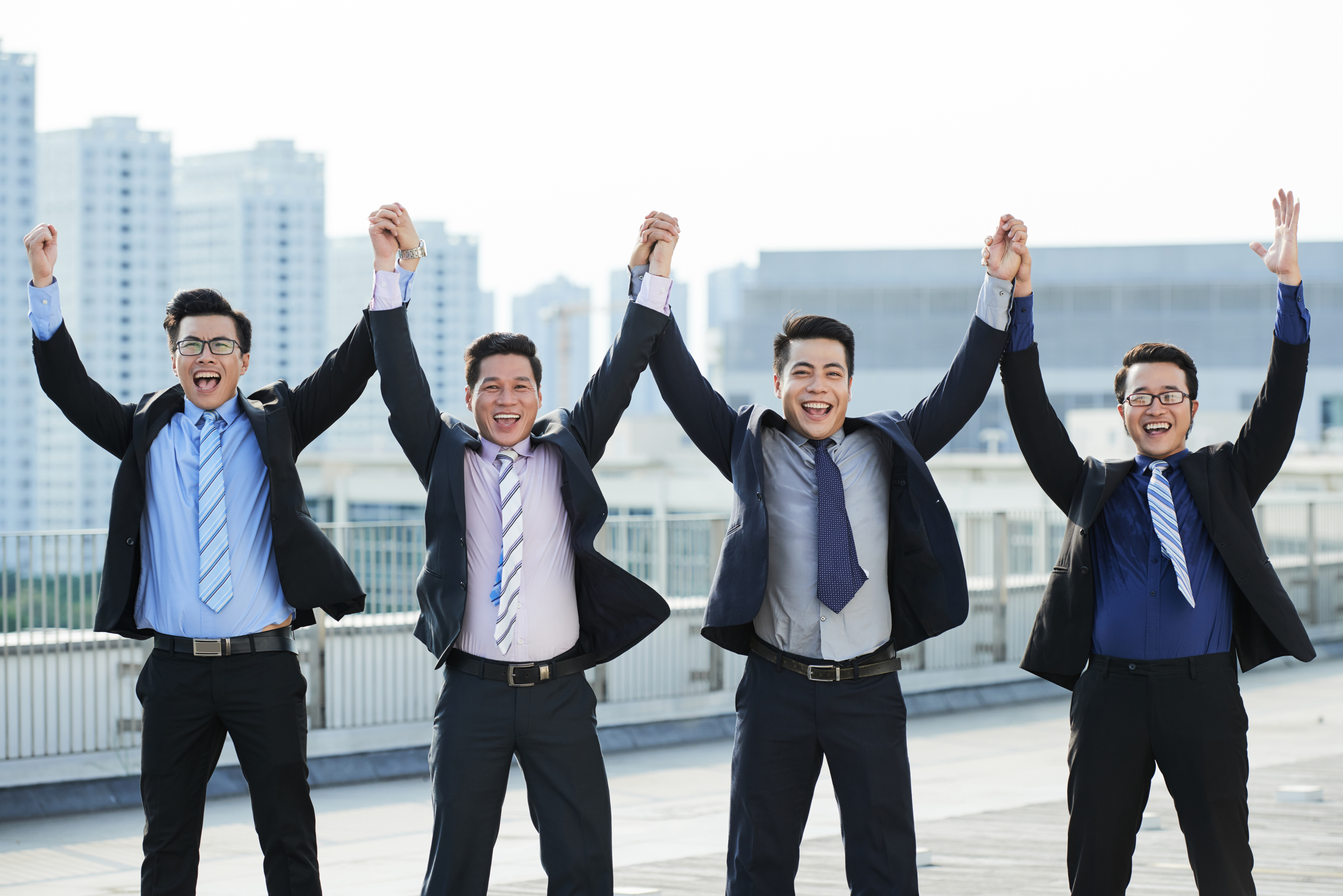 Cheerful white collar workers wearing suits holding hands and looking at camera with toothy smiles while celebrating successful completion of joint project, picturesque cityscape on background
